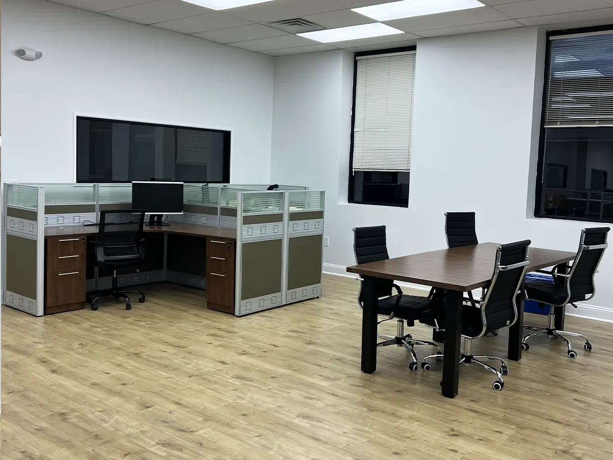 A cozy office corner with cubicle partitions, a desk with a monitor, and a meeting table surrounded by leather chairs.